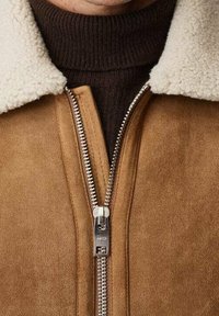 Ruskindsjakke i lysebrun med et krave af lammeskind. Har en sølvlynlås foran og en struktureret strikket underdel i mørkebrun.