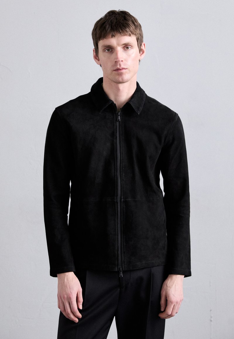 Veste en daim noir avec fermeture éclair à l'avant, col classique et manches longues. Elle présente une texture lisse et une silhouette ajustée.