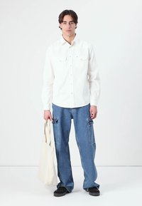 Hvit langermet skjorte med lommer foran, løse blå cargo-jeans med sidelommer, og svarte sko. En beige totebag holdes i den ene hånden.