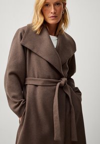Cappotto avvolgente in misto lana marrone con ampio colletto, vita regolabile con cintura e maniche lunghe. Texture liscia e design senza cuciture.