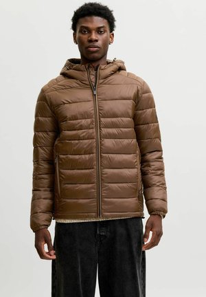 Jeune homme portant une veste matelassée marron à capuche avec fermeture éclair devant et un pantalon noir sur un fond clair uni.