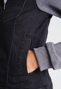 Svart denimjacka med grå sweatshirtärmar, framficka och textilstruktur. Kontrasterande sömmar synliga längs sömmarna.