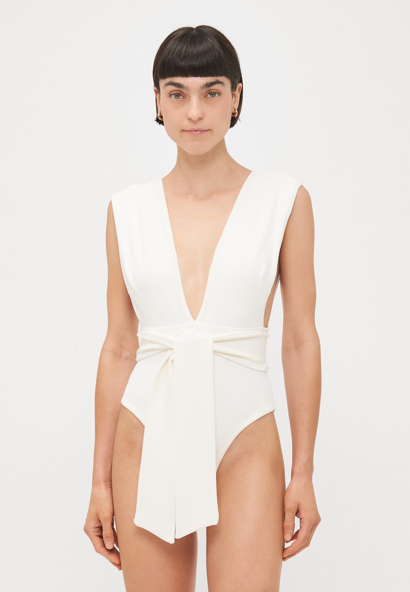 Un maillot de bain une pièce blanc avec un décolleté en V profond, des bretelles larges et un lien en tissu doux à la taille ; texture lisse et design ajusté.