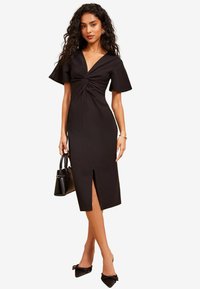 Schwarzes, tailliertes Kleid mit kurzen Ärmeln, einem verdrehten Knotendetail an der Taille, einem tiefen V-Ausschnitt und einem bis zum Oberschenkel reichenden Schlitz.