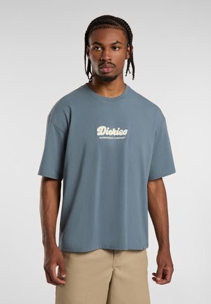 Camiseta de algodón azul claro con un corte holgado, que presenta un logo blanco "Dickies Workwear Company" en el pecho y mangas cortas.