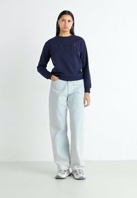 Sweatshirt azul-marinho com "PARIS" em letras em relevo, combinado com jeans de perna larga azul-claro e ténis cinzentos, sobre um fundo simples.
