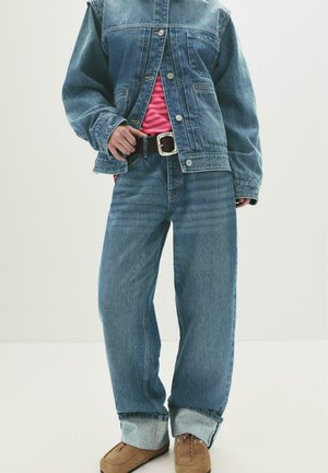 Person som bär en blå denimjacka och vida jeans över en rosa randig skjorta, med bruna mockaskor och ett svart bälte.