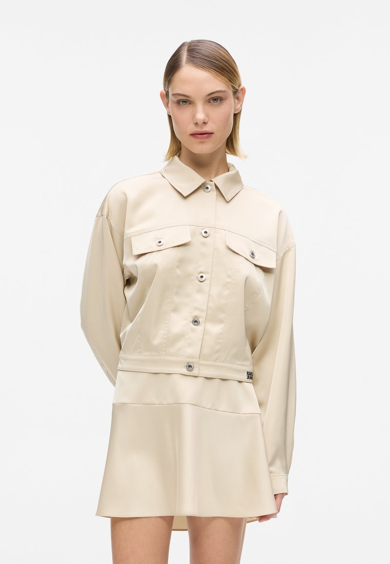 Beige Jacke mit Kragen, langen Ärmeln und zwei Brusttaschen. Glatter, leicht glänzender Stoff mit vorderen Druckknöpfen und einer zusätzlichen Rocklage.