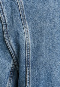 Světle modrá denimová tkanina s viditelným švem. Textura je mírně vybledlá, ukazuje hladký povrch s jemným vzorem tkaní.