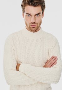Sweter w kolorze kremowym z warkoczy, z wysokim kołnierzem, z różnymi fakturowanymi wzorami i ściągaczami na mankietach. Długie rękawy o luźnym kroju.