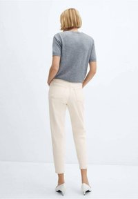 Femme aux cheveux blonds courts, vue de dos, portant un haut gris à manches courtes, un pantalon crème et des chaussures blanches à talons sur un fond blanc uni.