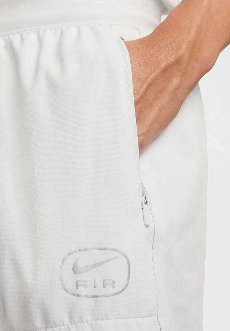 Shorts de sport blancs avec une poche zippée sur le côté et un logo Nike Air métallique discret sur la jambe gauche. Texture de tissu lisse.