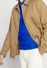 Vaaleanruskea takki, jossa on nappikiinnitys ja rintatasku, päällä sininen paita. Valkoiset housut täydentävät asustusta.