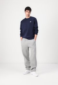 Navy crewneck sweatshirt met een wit logo, gecombineerd met lichtgrijze sweatpants en witte sneakers. Minimalistisch ontwerp, relaxed fit.