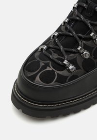 Schwarzer Wanderschuh mit einem gemusterten Obermaterial, das dunkelgraue Akzente aufweist. Metallische Ösen stützen die dicken Schnürsenkel; robuste Sohle für besseren Grip.