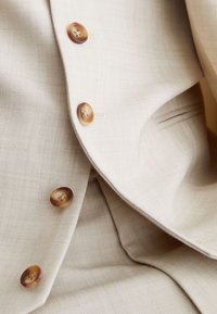 Beige tekstureret stof med skildpaddemønstrede knapper på en skræddersyet jakke eller frakke med fokus på knappens detaljer og reversets fold.