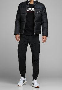 Chaqueta acolchada negra con cuello alto, detalle de cremallera, combinada con una camiseta negra con texto blanco, pantalones cargo negros y zapatillas blancas.