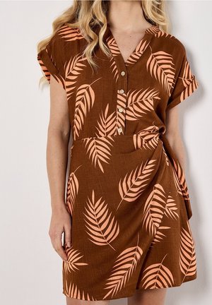 Femme portant une robe marron à manches courtes avec des motifs de feuilles de pêche, un col en V et des boutons sur le devant, debout devant un fond blanc.