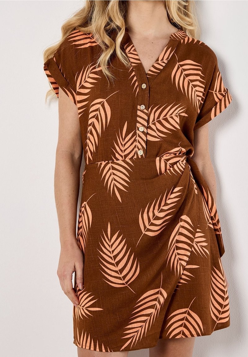 Femme portant une robe marron à manches courtes avec des motifs de feuilles de pêche, un col en V et des boutons sur le devant, debout devant un fond blanc.