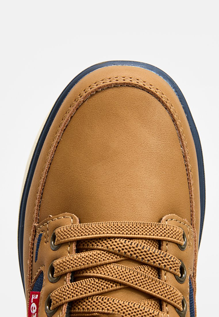 Gros plan sur l'orteil d'une botte en cuir beige avec des coutures détaillées, des lacets tissés marron, des œillets en métal et des accents bleu marine le long de la semelle et du côté.