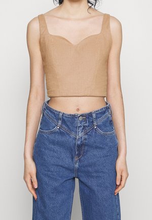 Vrouw die een mouwloos beige crop top en een blauwe high-waisted spijkerbroek met knoop- en ritssluiting draagt.