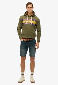 Sudadera verde oliva con cordones blancos y una franja de logo, llevada sobre una camisa de cuadros rojos. Combinada con unos pantalones cortos de mezclilla azules y zapatillas beige.