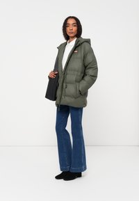 Maslinasto zelena jakna s kapuljačom, sužena u struku, s prednjim džepovima i logotipom. Upari sa plavim zvonastim trapericama i crnim čizmama do gležnja.