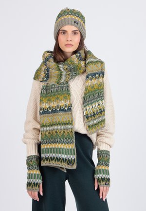 Frau trägt einen beigen Pullover, eine grüne gemusterte Strickmütze, einen passenden Schal und fingerlose Handschuhe, steht vor einem weißen Hintergrund.