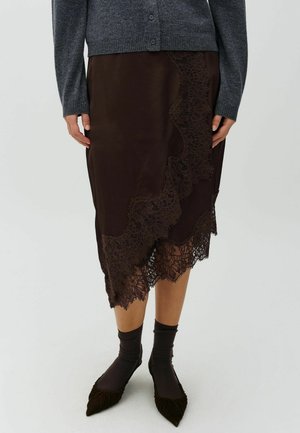 Woman wearing a dark brown lace-trimmed midi skirt, dark socks, pointed shoes, and a gray buttoned cardigan.