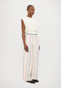 Teksturojana bela pletena majica s kratkimi rokavi v kombinaciji z širokimi trakastimi hlačami v pastelnih barvah, ki imajo vertikalne črte in ohlapen kroj.