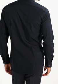 Camisa negra de manga larga hecha de tejido de algodón suave, con cuello puntiagudo, puños con botones y bajo curvado.