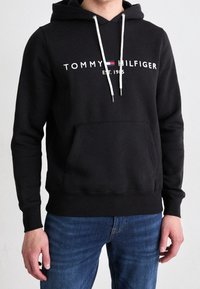 Man som bär en svart Tommy Hilfiger hoodie med vita dragsko och logotyp, tillsammans med blå jeans, står mot en neutral bakgrund.