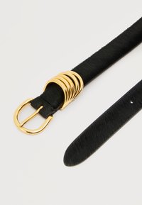 Cinturón negro texturizado con hebilla dorada y cinco anillas doradas, colocado plano sobre una superficie blanca.