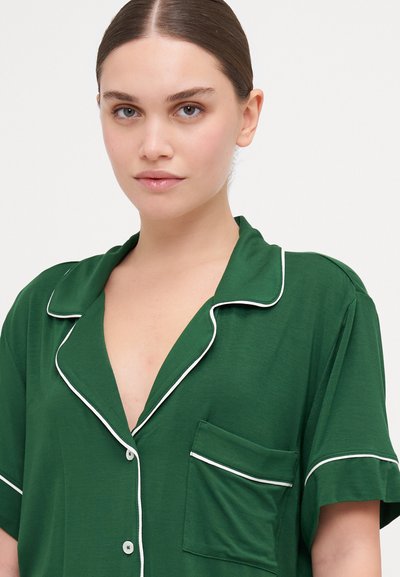 Camisa de pijama verde de manga curta feita de tecido macio, com colarinho picotado, ribete branco e um único bolso no peito.