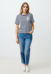 Marineblauw en witte gestreepte t-shirt met korte mouwen en ronde halslijn, voorzien van een klein grafisch detail. Gecombineerd met lichtblauwe rechte jeans en witte sneakers.