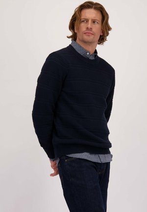 Marineblauwe gebreide trui met horizontale strepen, ronde halslijn, gedragen over een lichtblauwe overhemd met kraag, gecombineerd met donkere jeans.