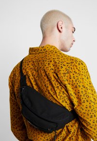 Svart stoff crossover-veske med en glidelåslomme, båret over en gul skjorte med et svart leopardmønster.