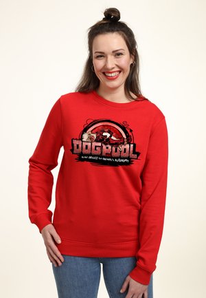 Woman with dark hair in a bun smiling, wearing a red sweatshirt with a "Dogpool" graphic combining a dog and superhero theme.