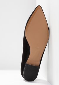 Chaussure à talon haut en daim noir avec un bout pointu et une semelle beige, dotée d'un talon noir texturé. Positionnée debout sur un fond blanc.