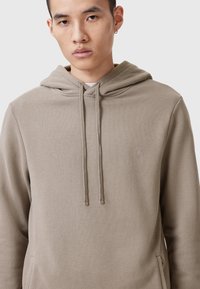 Sudadera beige con cordones y un bolsillo tipo canguro. Presenta un logotipo sutil en gris claro en el pecho, fabricada con un tejido suave y texturizado.