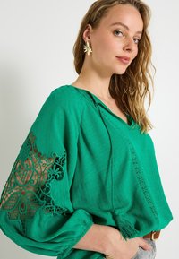 Blouse verte avec un col en V, en tissu texturé, et avec des détails en dentelleintriqués sur les manches. Présente des manches longues amples et une coupe décontractée.