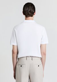 Weißes Poloshirt mit kurzen Ärmeln und strukturiertem Finish, von hinten gezeigt, kombiniert mit hellbeigen, gestreiften Hosen.