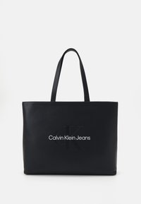 Imanska torba od crne kože s dvostrukim čvrstim ručkama, uvezene "CK" oznake i natpisom "Calvin Klein Jeans" ispisanim u bijelom. Glatke teksture, pravokutnog oblika.