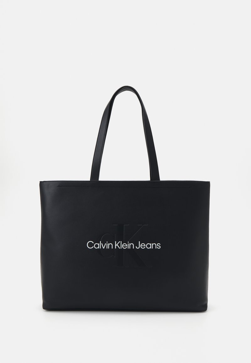 Imanska torba od crne kože s dvostrukim čvrstim ručkama, uvezene "CK" oznake i natpisom "Calvin Klein Jeans" ispisanim u bijelom. Glatke teksture, pravokutnog oblika.