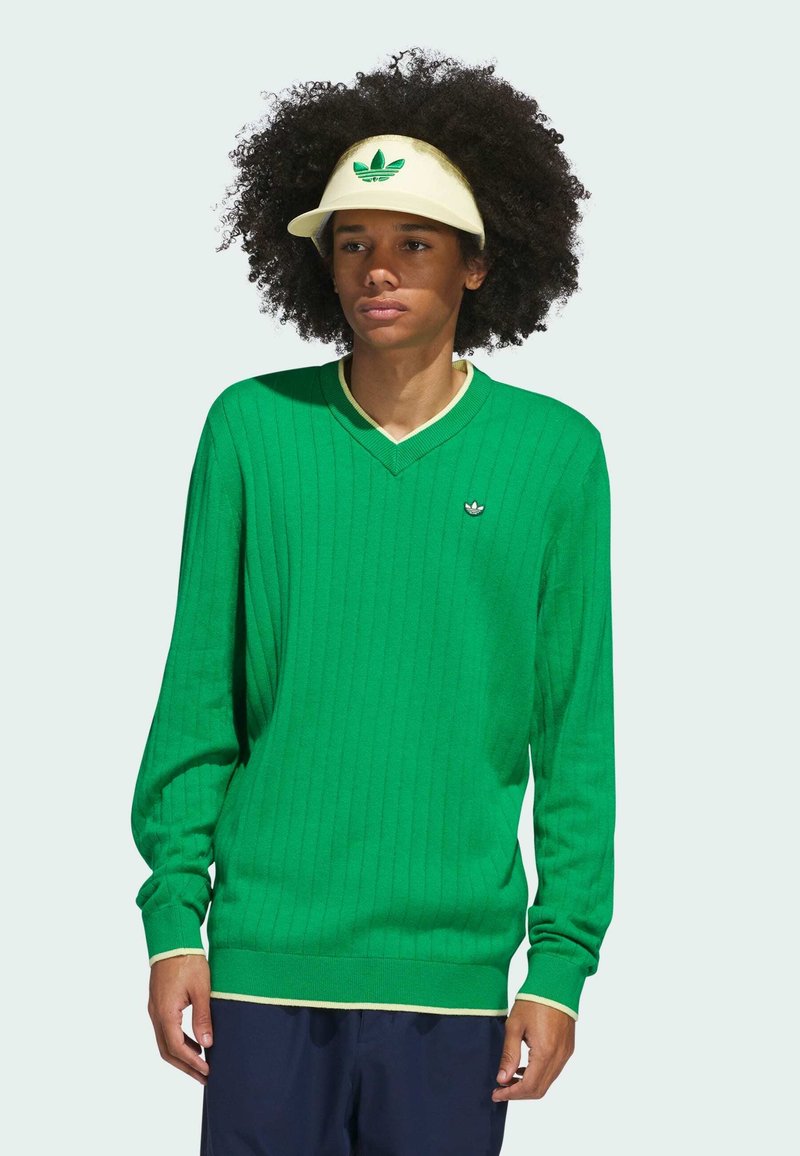 Grøn v-hals sweater med tekstureret ribning, cremede detaljer ved halsudskæringen og kanten, lille broderet logo på brystet. Modellen bærer en cremet skygge.