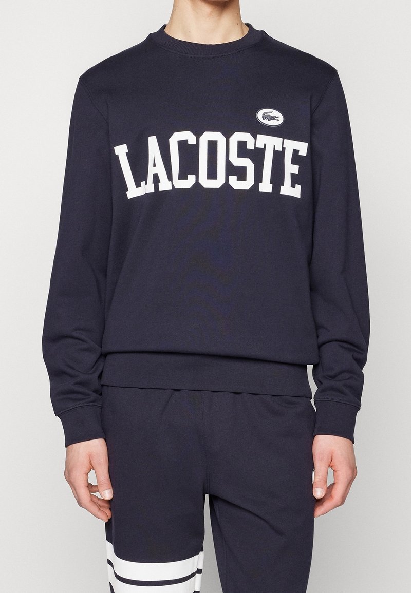 Mörkblå sweatshirt med vit "LACOSTE"-text och broderad logotyp. Har rundad halsringning, långa ärmar samt ribbade muddar och fåll.