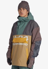 Bunte Kapuzenjacke mit grünem Oberteil, braunen Ärmeln und einer gelben Tasche. Hat auffällige weiße Markenkennzeichnung und einen strukturierten dunkleren Ärmel.