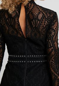 Vue de dos d'une personne portant une robe en dentelle noire avec une ouverture en forme de trou de serrure et deux ceintures ornées d'œillets à la taille.
