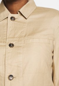 Giacca beige con colletto, quattro bottoni e tasca sul petto. Realizzata in un tessuto leggero con una texture liscia e finiture cucite.