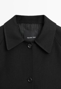 Cappotto nero con colletto classico, chiusura frontale con bottoni e interno foderato a righe. Le etichette sono visibili all'interno del colletto.